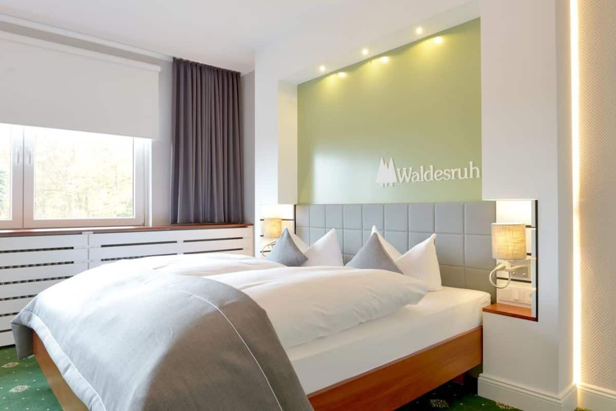 A hotel room with a large bed, a headboard with a logo, a window with curtains, and a radiator below the window.