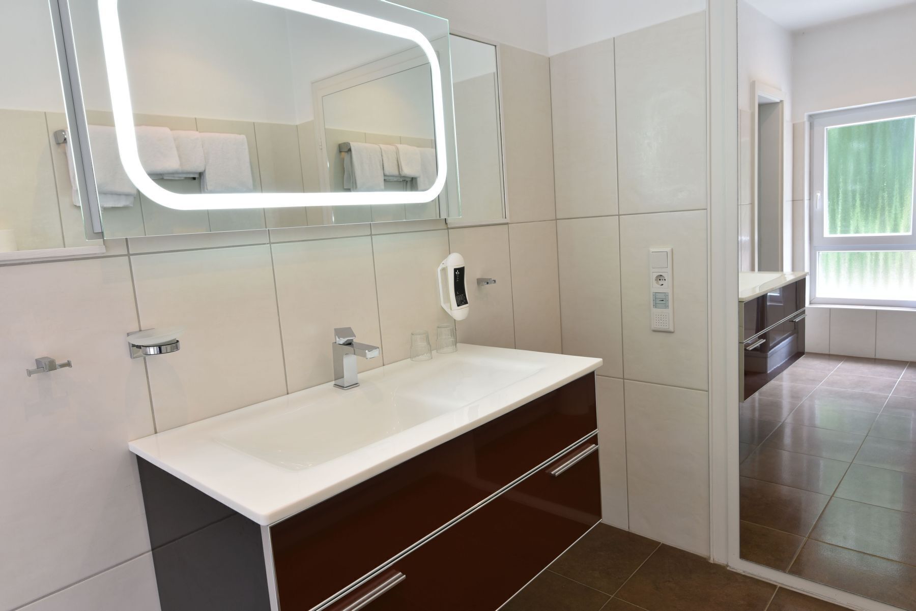 Un baño moderno con tocador blanco, un espejo con iluminación integrada, un perchero y un panel de control de ducha.