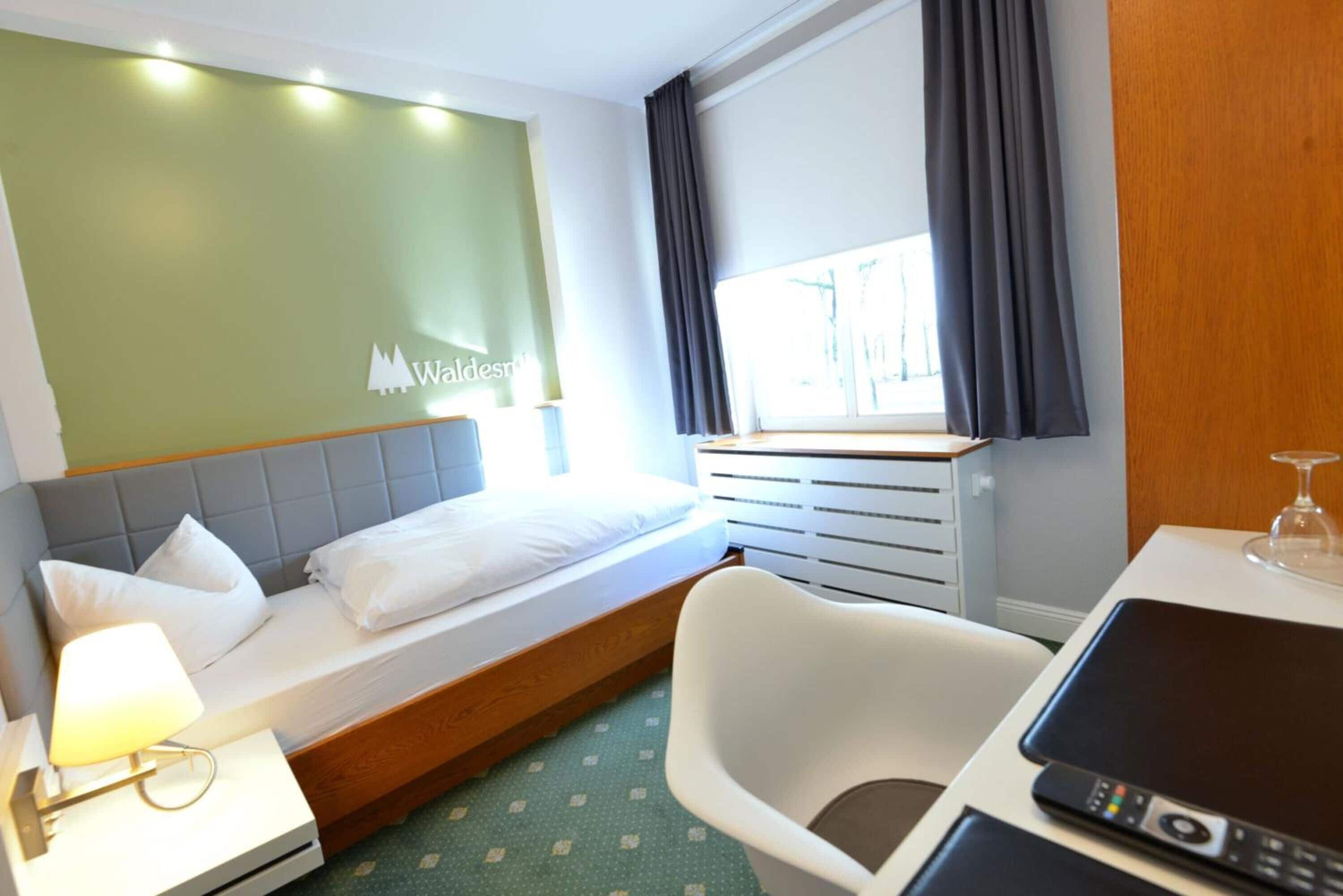 A hotel room with a bed, a desk with a lamp, a chair, and a window with curtains.