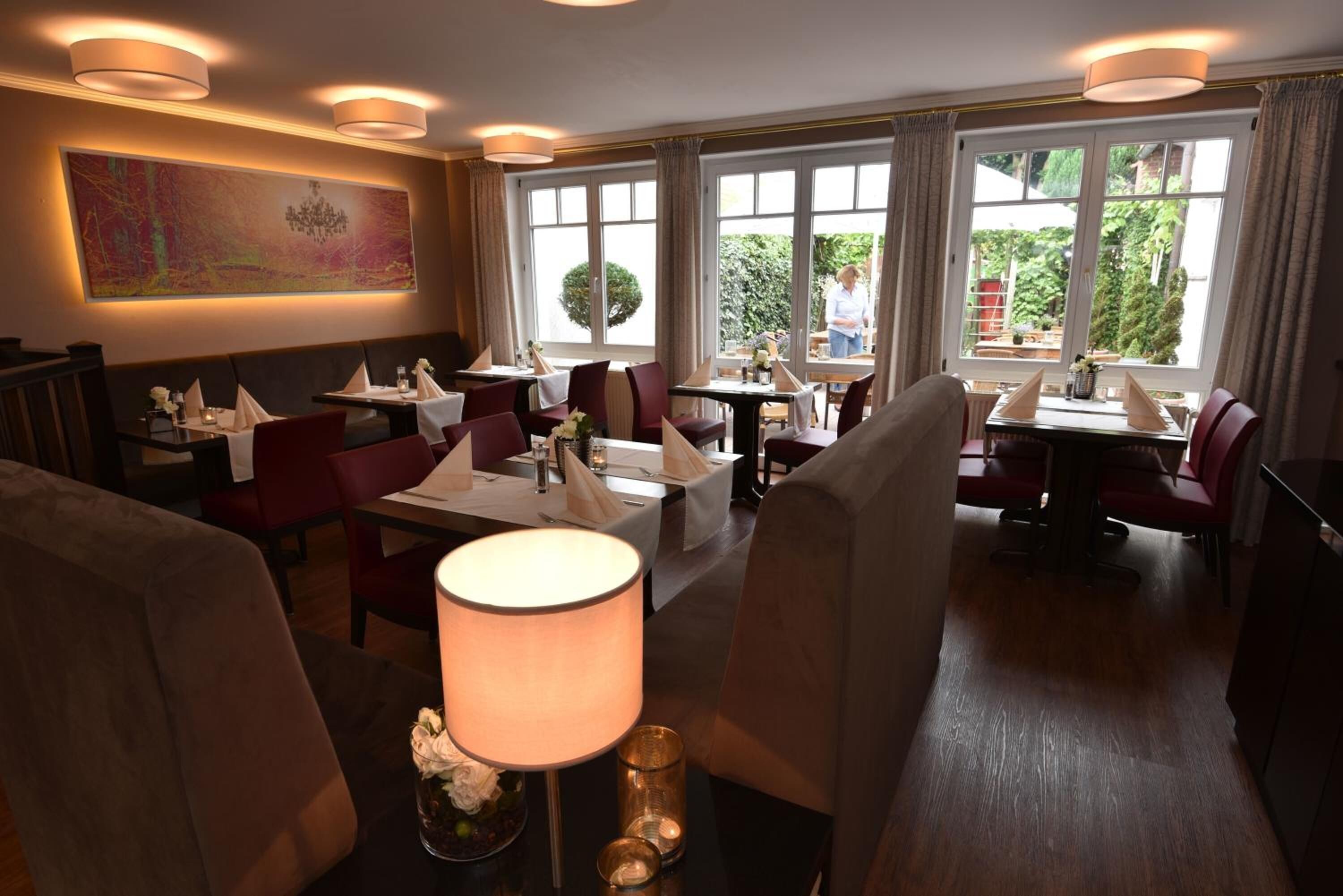 Un comedor con mesas dispuestas para dos, ventanas amplias con cortinas y un candelabro en la pared.