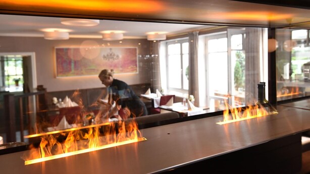 A restaurant with a modern interior, featuring a long counter with a grill and flames, and a view of the dining area through large windows. - Hotel Restaurant Waldesruh (Emstek)