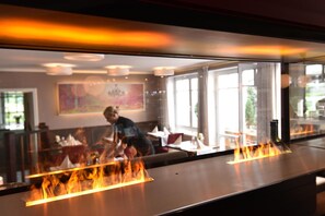 A restaurant with a modern interior, featuring a long counter with a grill and flames, and a view of the dining area through large windows. - Hotel Restaurant Waldesruh (Emstek)