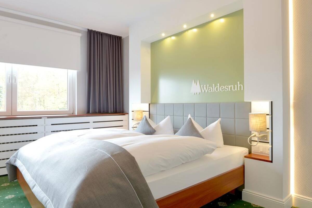 A hotel room with a large bed, a headboard with a logo, bedside lamps, and a window with curtains.