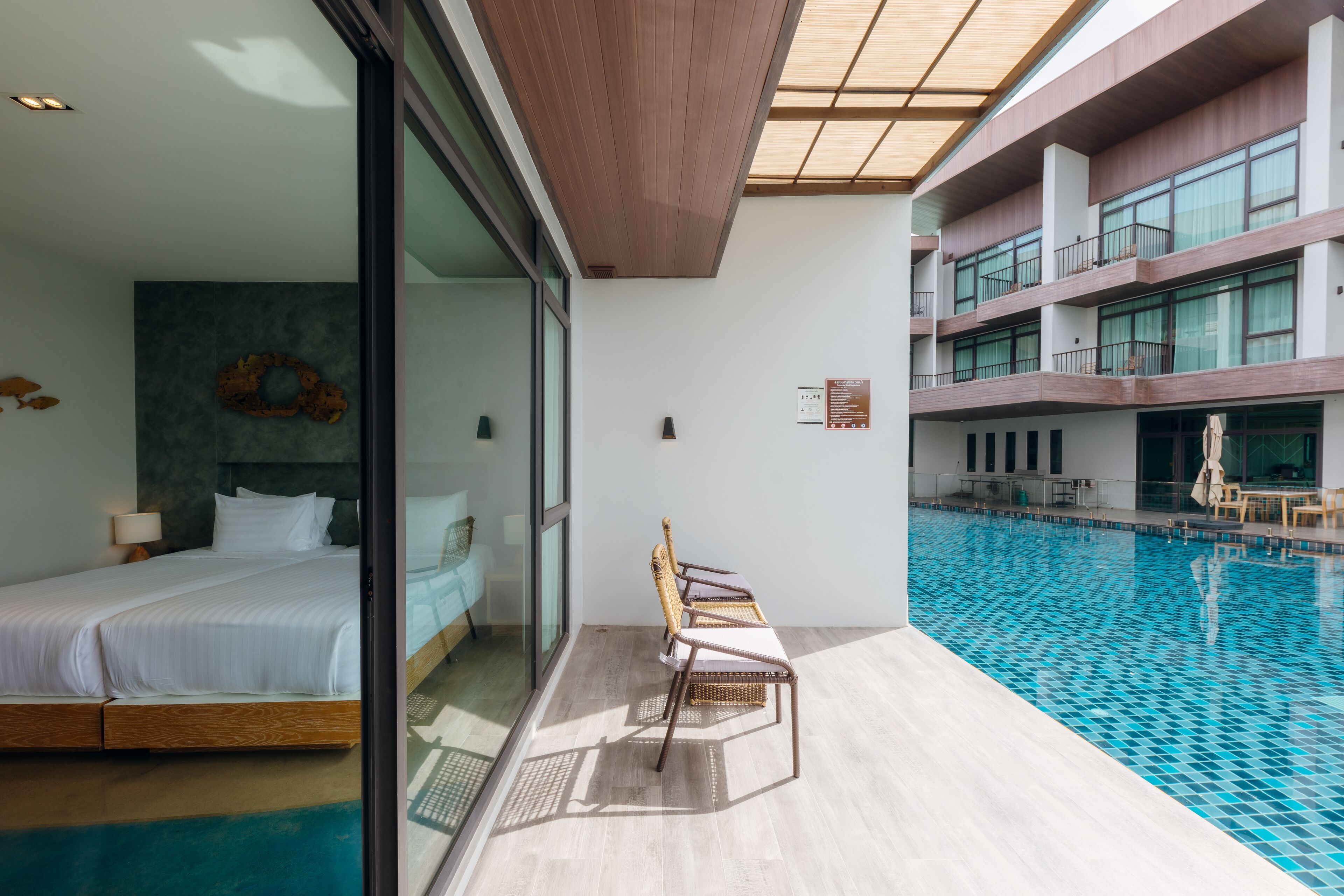 Una habitación de hotel moderna con una cama grande, un muro de cristal, una piscina y un balcón con sillas de mimbre.