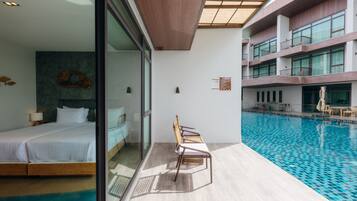 Una habitación de hotel moderna con una cama grande, un muro de cristal, una piscina y un balcón con sillas de mimbre.