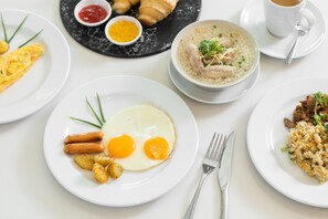 A breakfast setting with plates of eggs, sausages, and fried potatoes, a bowl of rice with meat, and croissants.
