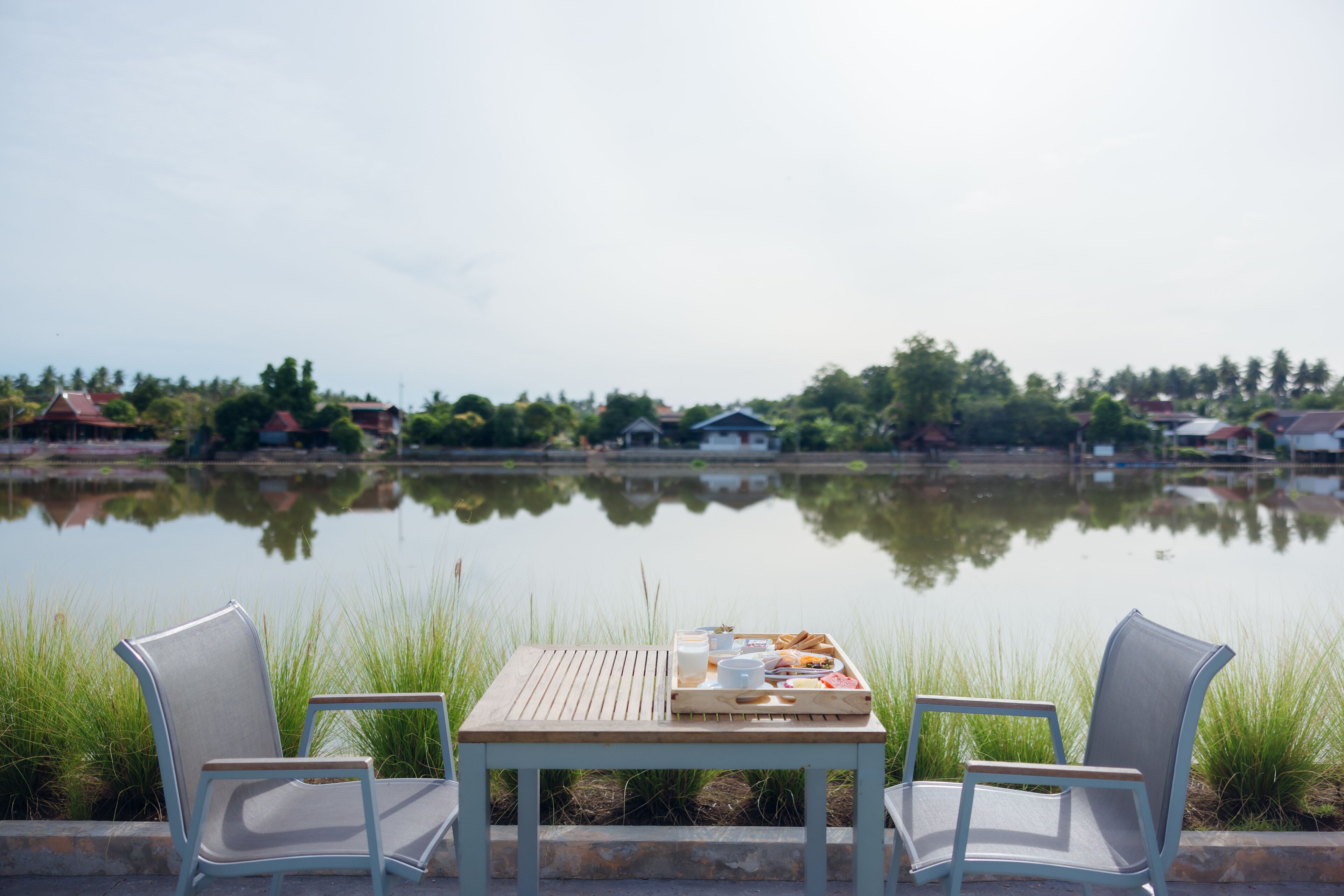 Una mesa con una bandeja de comida y bebidas, dos sillas, y vista a un cuerpo de agua tranquilo con casas al fondo.