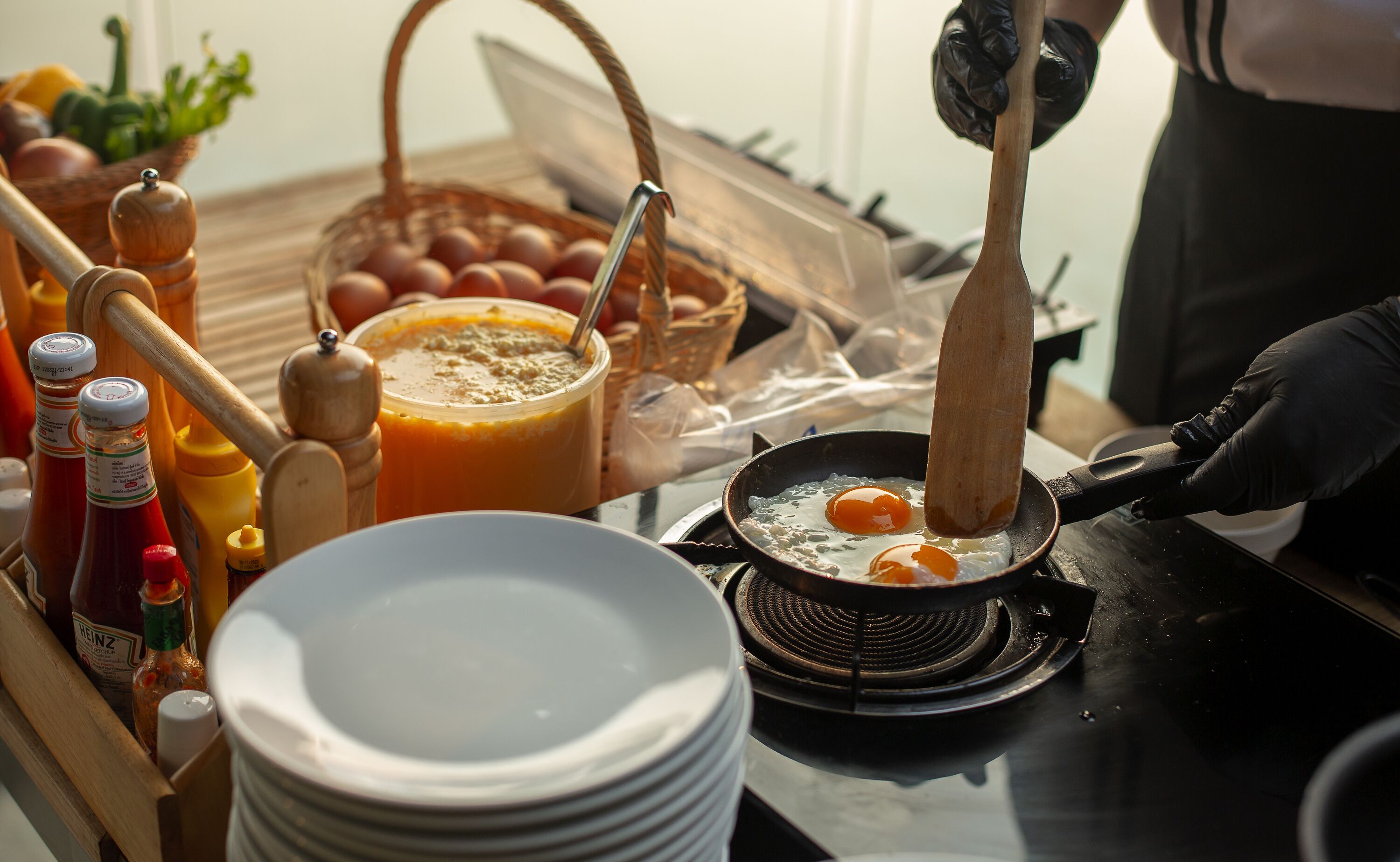A breakfast setup with eggs being fried, condiments, and a basket of eggs on a table.