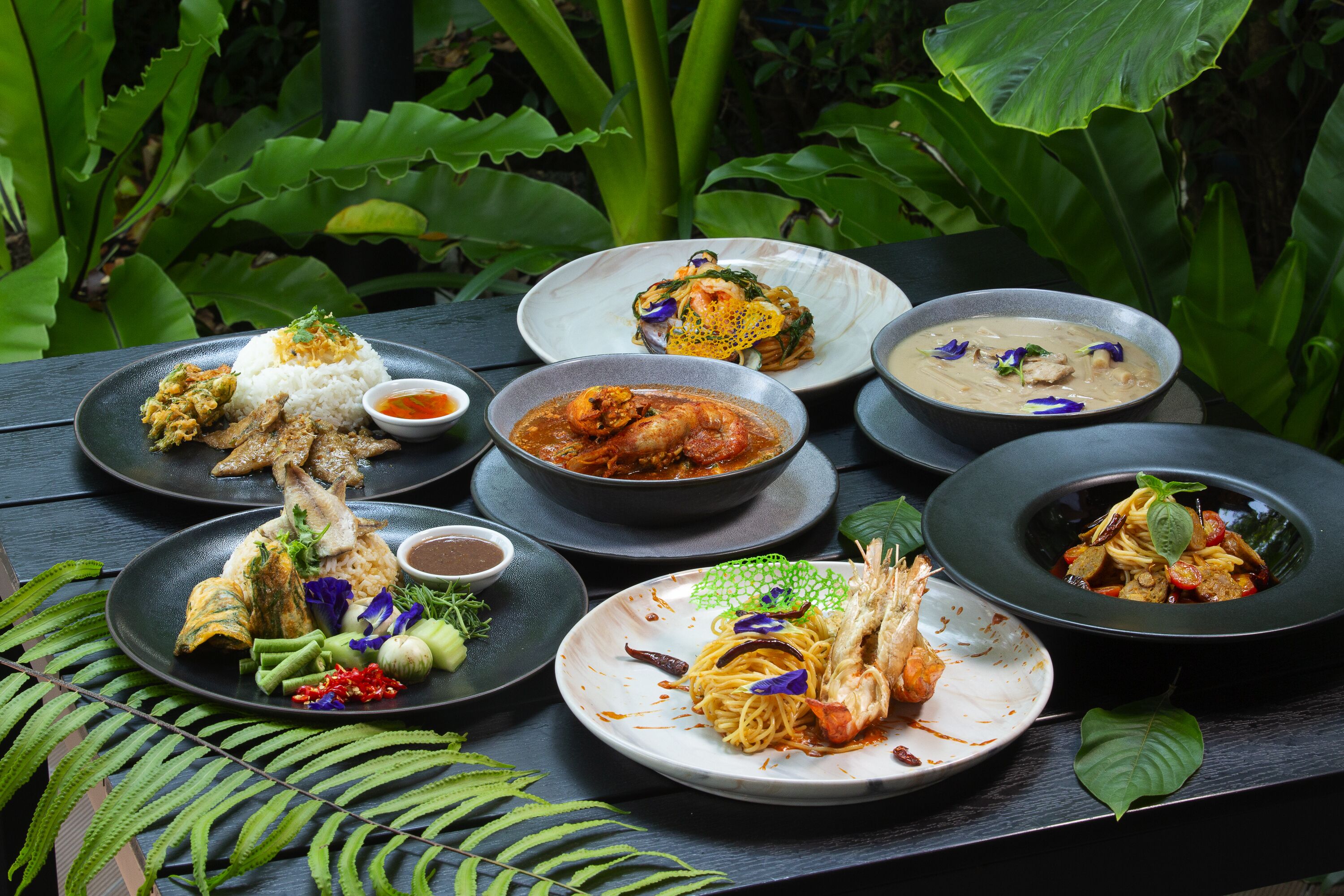 A table set with various dishes, including rice, seafood, and curry, surrounded by lush green foliage.