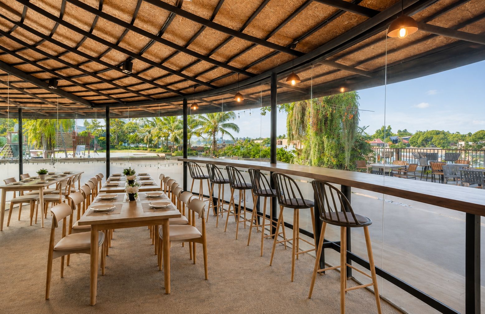 Un área de comedor al aire libre con mesas y sillas, techo de paja y vista a palmeras y edificios.