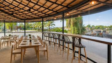 Un área de comedor al aire libre con mesas y sillas, techo de paja y vista a palmeras y edificios.