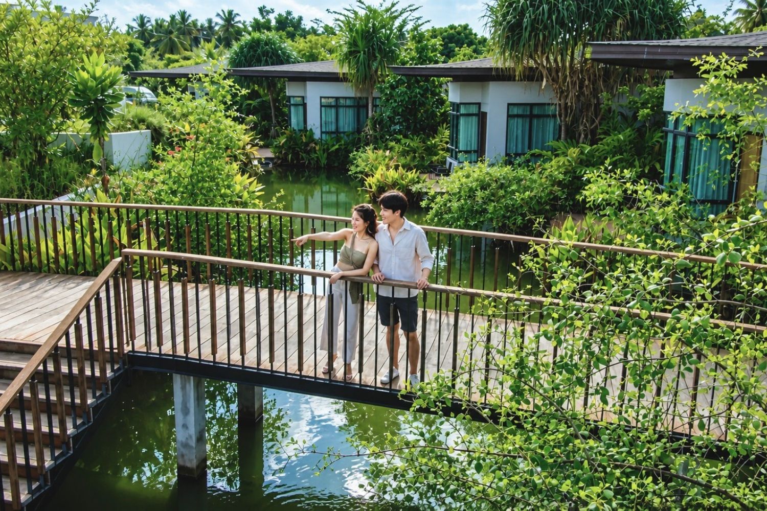 A wooden bridge over a body of water with lush greenery and modern buildings in the background.