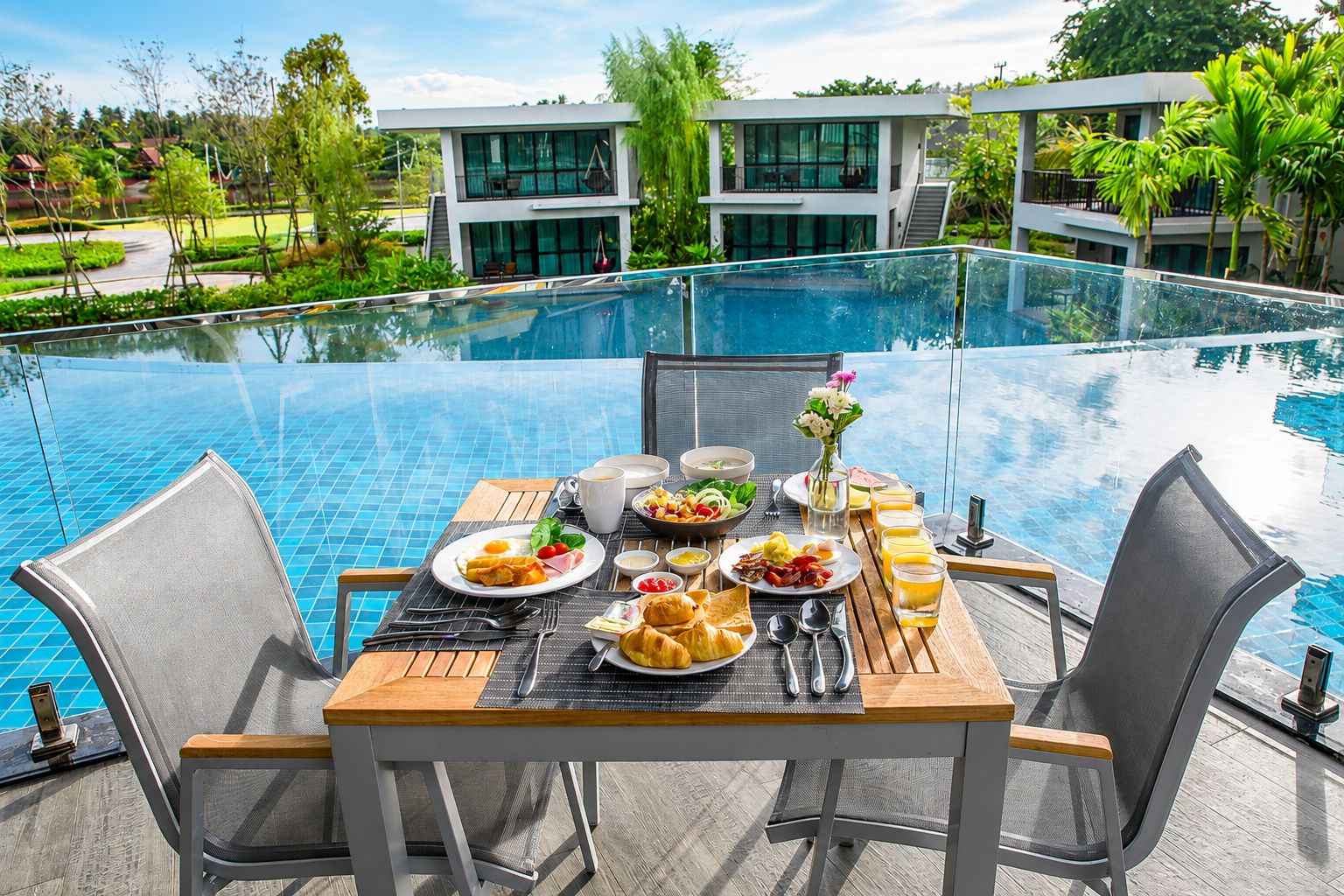Mesa de comedor con desayuno en una terraza junto a la piscina, con vista a un edificio moderno y exuberante vegetación.
