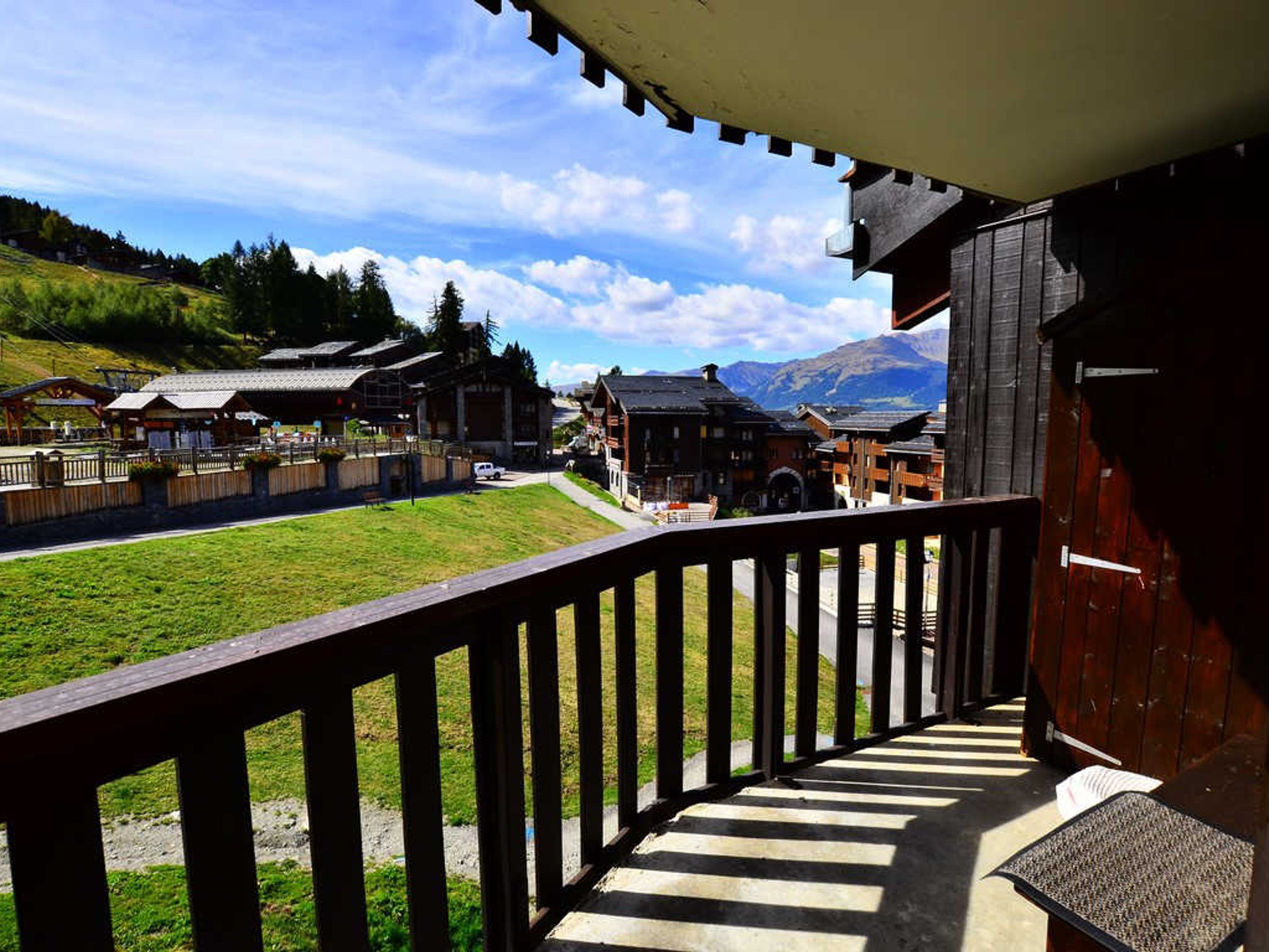 A wooden balcony overlooking a village with traditional buildings, a grassy area, and mountains in the background.