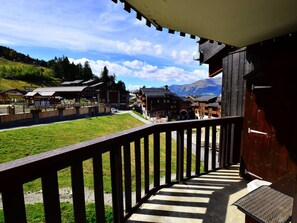 A wooden balcony overlooking a village with traditional buildings, a grassy area, and mountains in the background.