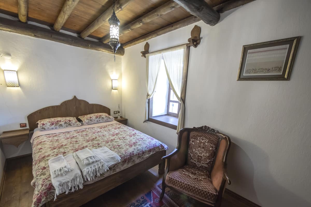Una camera da letto con un letto in legno, un comodino, una sedia, una finestra con tende e un punto luce a parete.