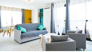 Een moderne woonkamer met een grijze bank, kussens in teal-kleur, een witte salontafel en grijze fauteuils. Er is een eethoek met een witte tafel en stoelen, en een balkon met uitzicht.