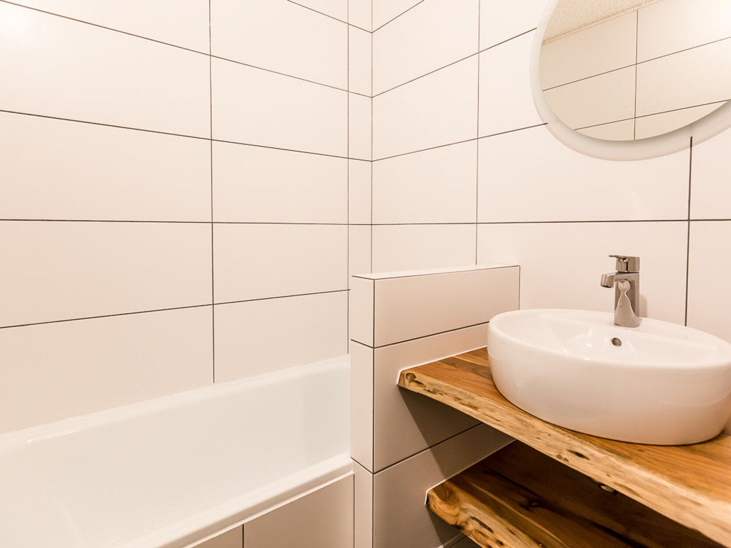 Une salle de bain avec des carreaux blancs, un meuble-lavabo en bois, un miroir rond et un lavabo blanc.