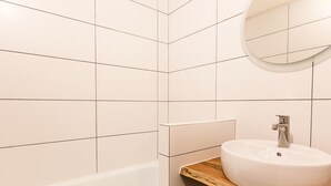 A bathroom with white tiles, a wooden vanity, a round mirror, and a white sink. - Divisible studio for 3, near shops and slopes, free parking, balcony (Bellentre)