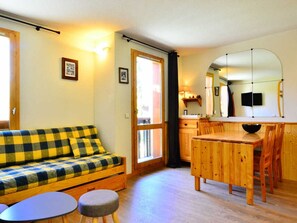 A cozy living room with a checkered sofa, a small round table, and a wooden dining table with chairs. There is a kitchen area with a refrigerator, a sink, and a mirror.