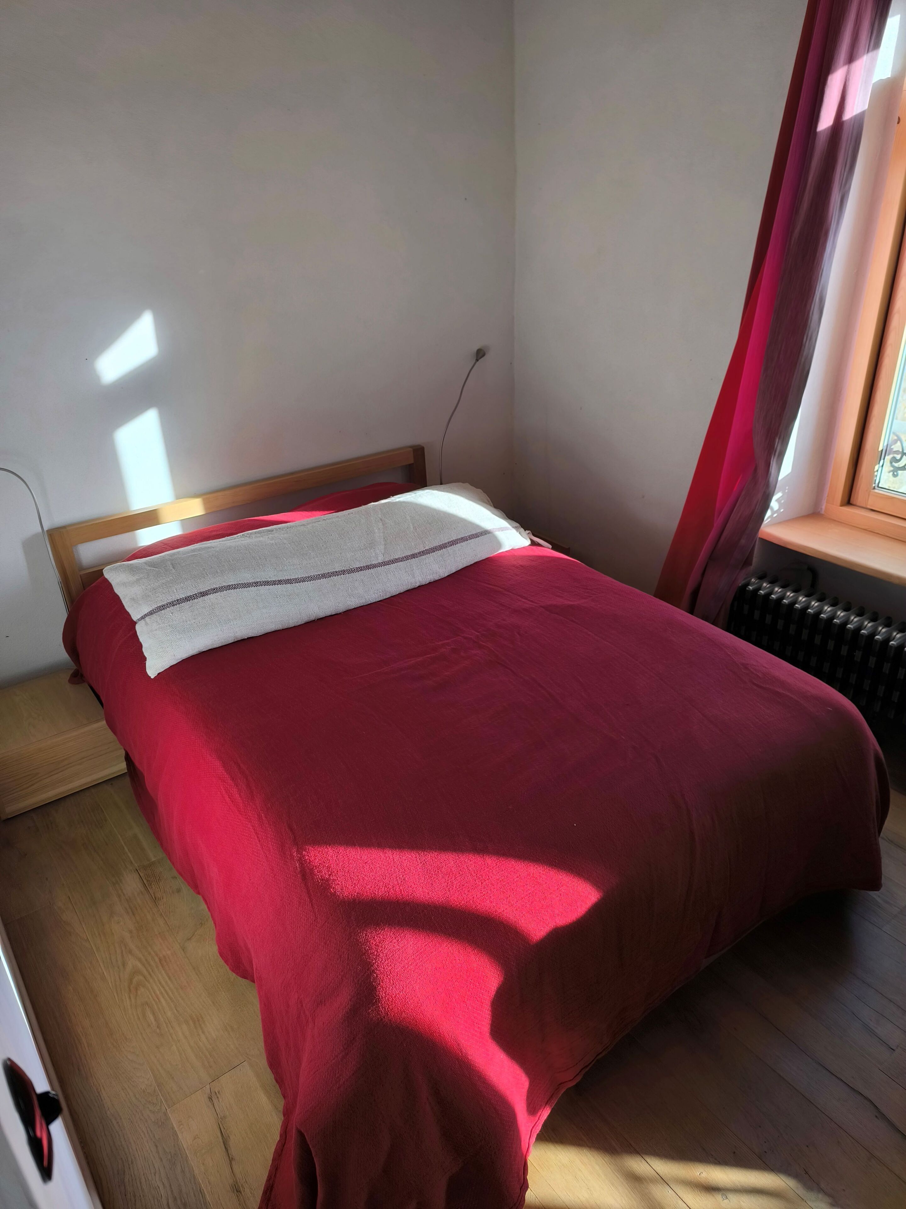 Een eenpersoonsbed met een rode sprei en een wit kussen in een kamer met houten vloeren en een radiator.