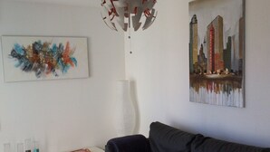 A modern living room with a dark grey sofa, a white side table with a framed picture, a colorful abstract painting, and a cityscape painting on the wall. - T2 Very quiet, close to beaches and town center, sea and mountain views (L'île-Rousse)