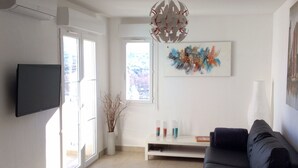 A modern living room with a flat-screen TV, a black sofa, a decorative ceiling light, a window with a view, and a colorful abstract painting on the wall. - T2 Very quiet, close to beaches and town center, sea and mountain views (L'île-Rousse)