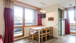 A dining area with a table and chairs, large windows with curtains, a television mounted on the wall, and a green door. - Apartment for 6 with balcony, ski lockers, near slopes and shops (Bellentre)