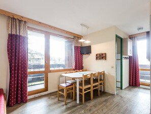 A dining area with a table and chairs, large windows with curtains, a television mounted on the wall, and a green door.
