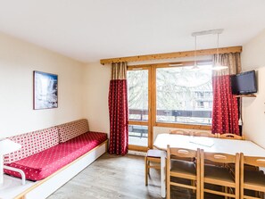 A cozy room with a wooden bench, a dining table with chairs, a television mounted on the wall, and a window with curtains.