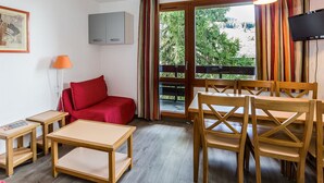 Ein Zimmer mit Holztisch und Stühlen, einem roten Sofa, einer Lampe, einem Fernseher und einem Fenster mit Vorhängen.