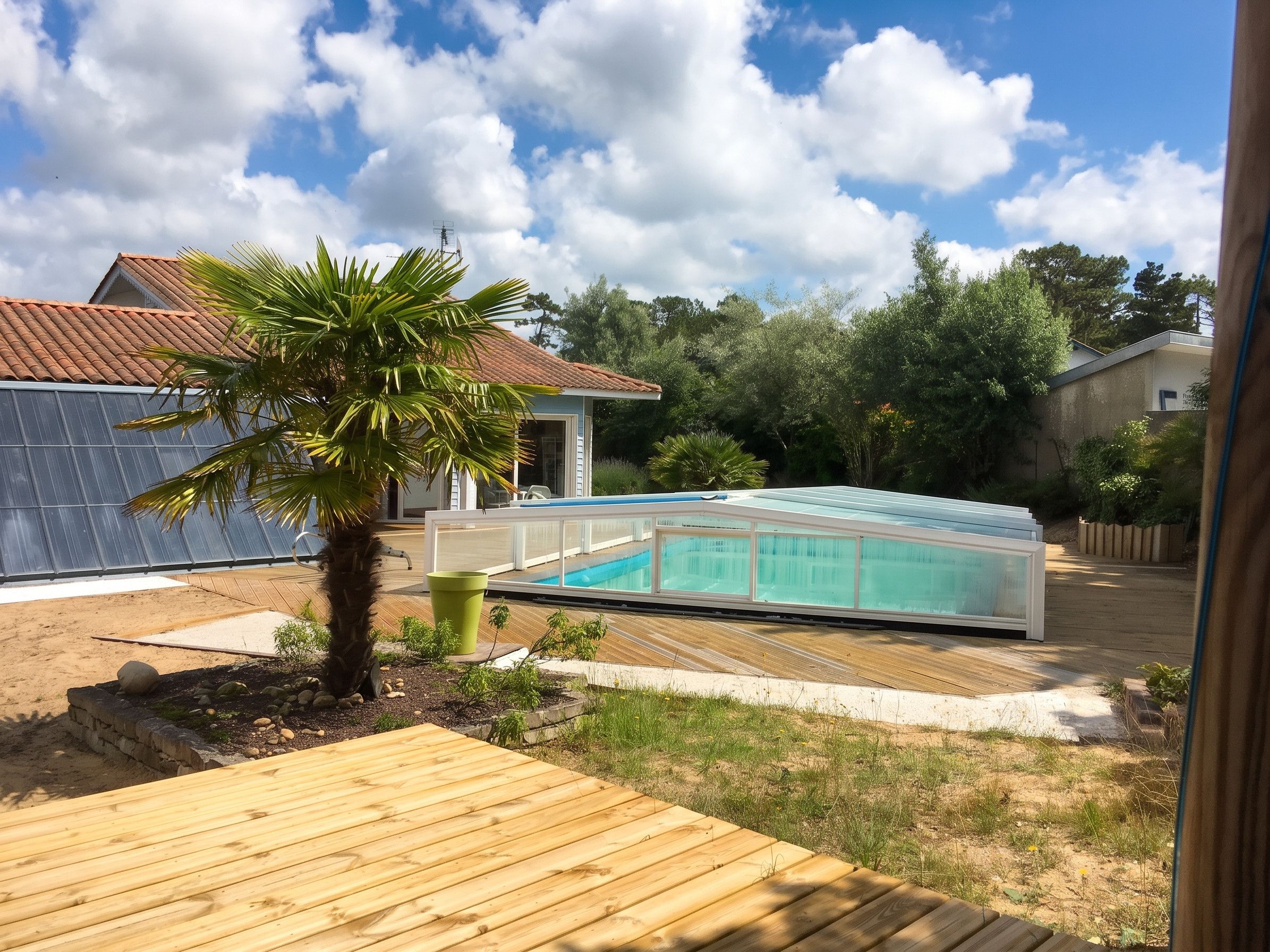 A swimming pool with a glass enclosure, surrounded by a wooden deck, a palm tree, and a house with a tiled roof.