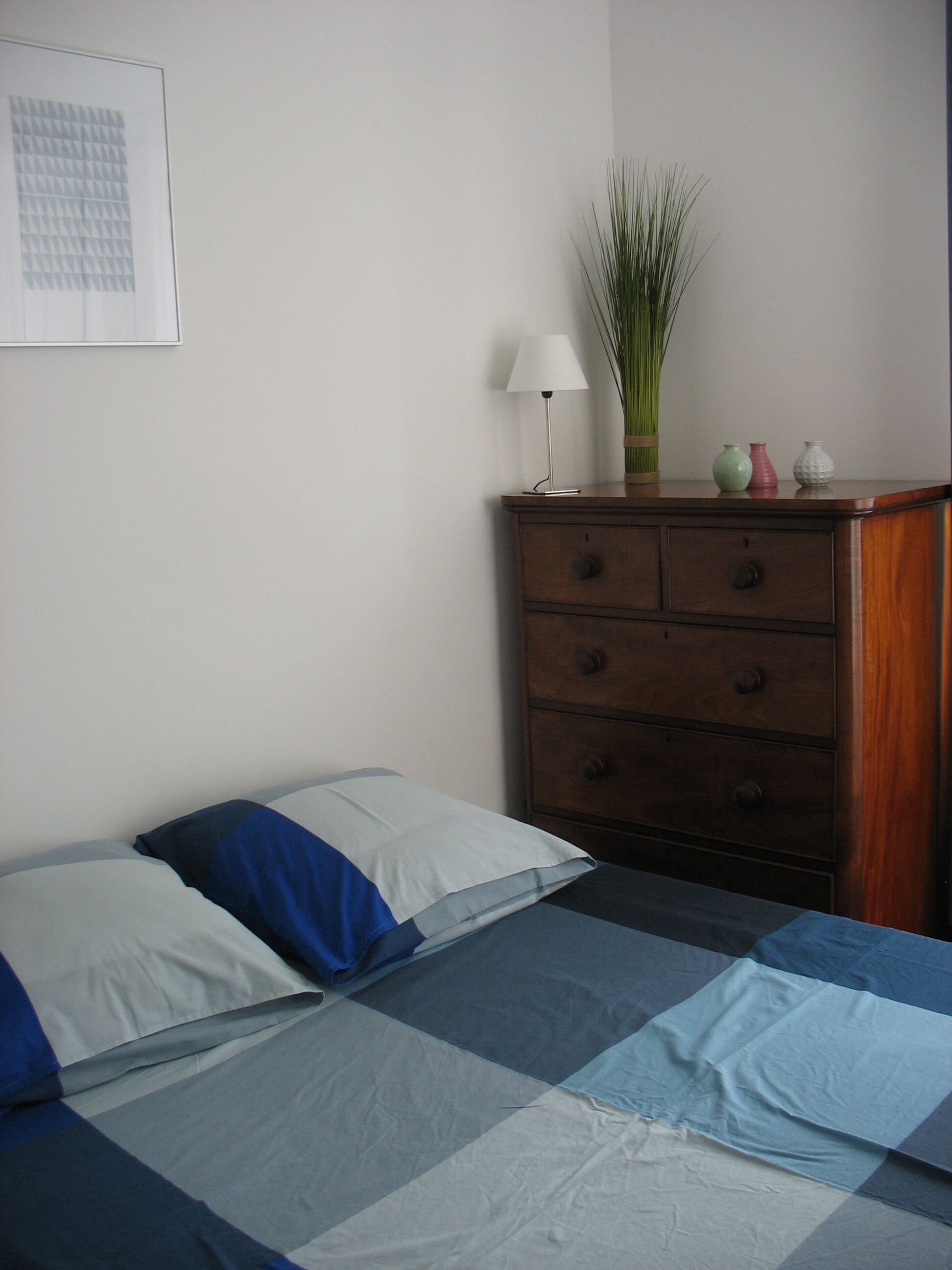 Une chambre à coucher avec un lit, deux oreillers, une commode, une lampe et une plante.