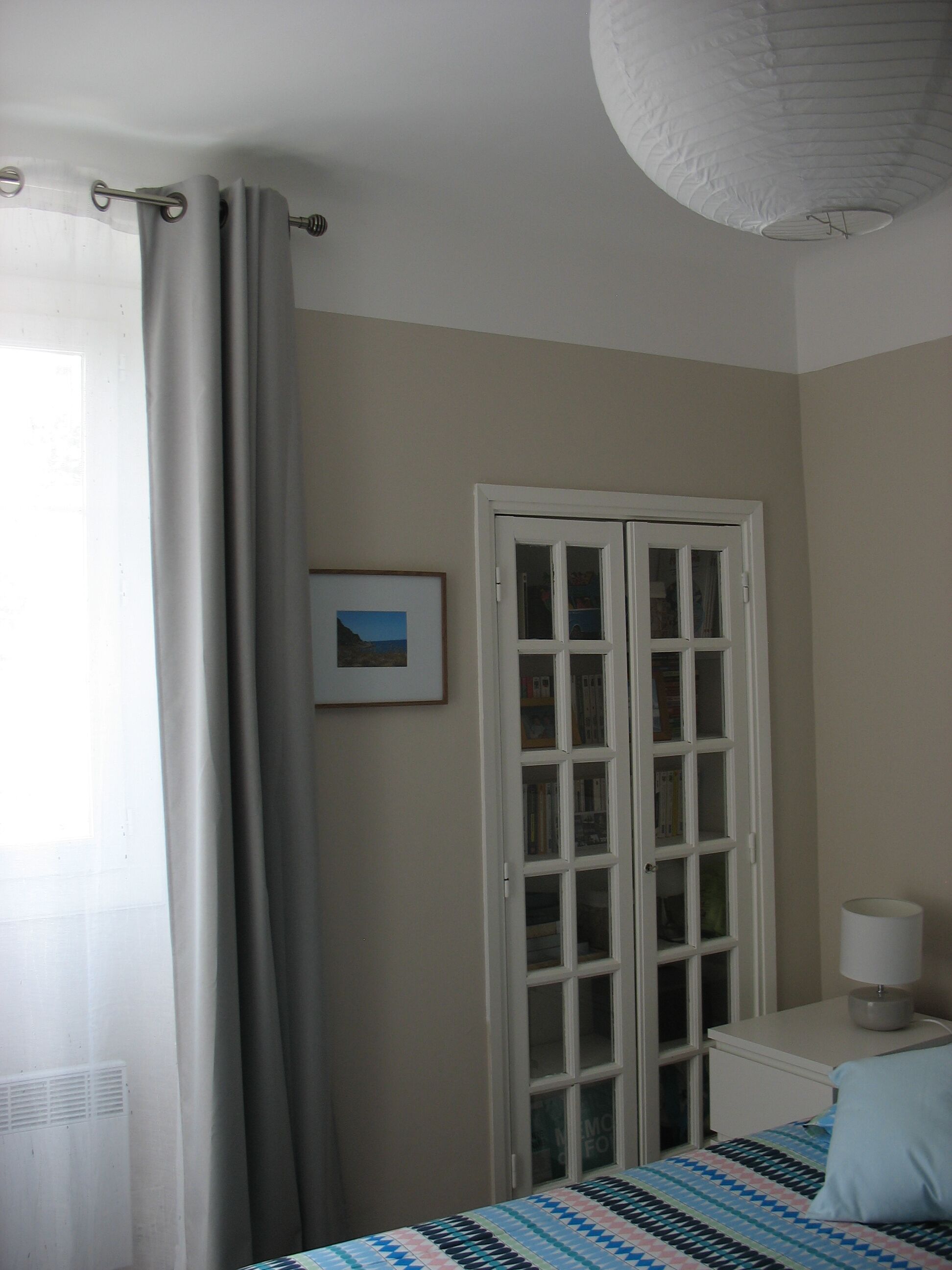 Une chambre à coucher comprenant un lit, une table de chevet, une lampe, une porte avec des panneaux de verre et une fenêtre avec des rideaux.