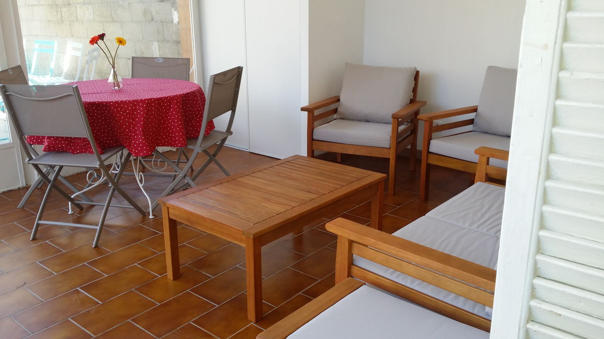 A patio area with a wooden table, two cushioned chairs, and a red tablecloth.