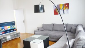 Ein modernes Wohnzimmer mit einem grauen Ecksofa, einem Glastisch und einem Flachbildfernseher auf einem Standfuß.