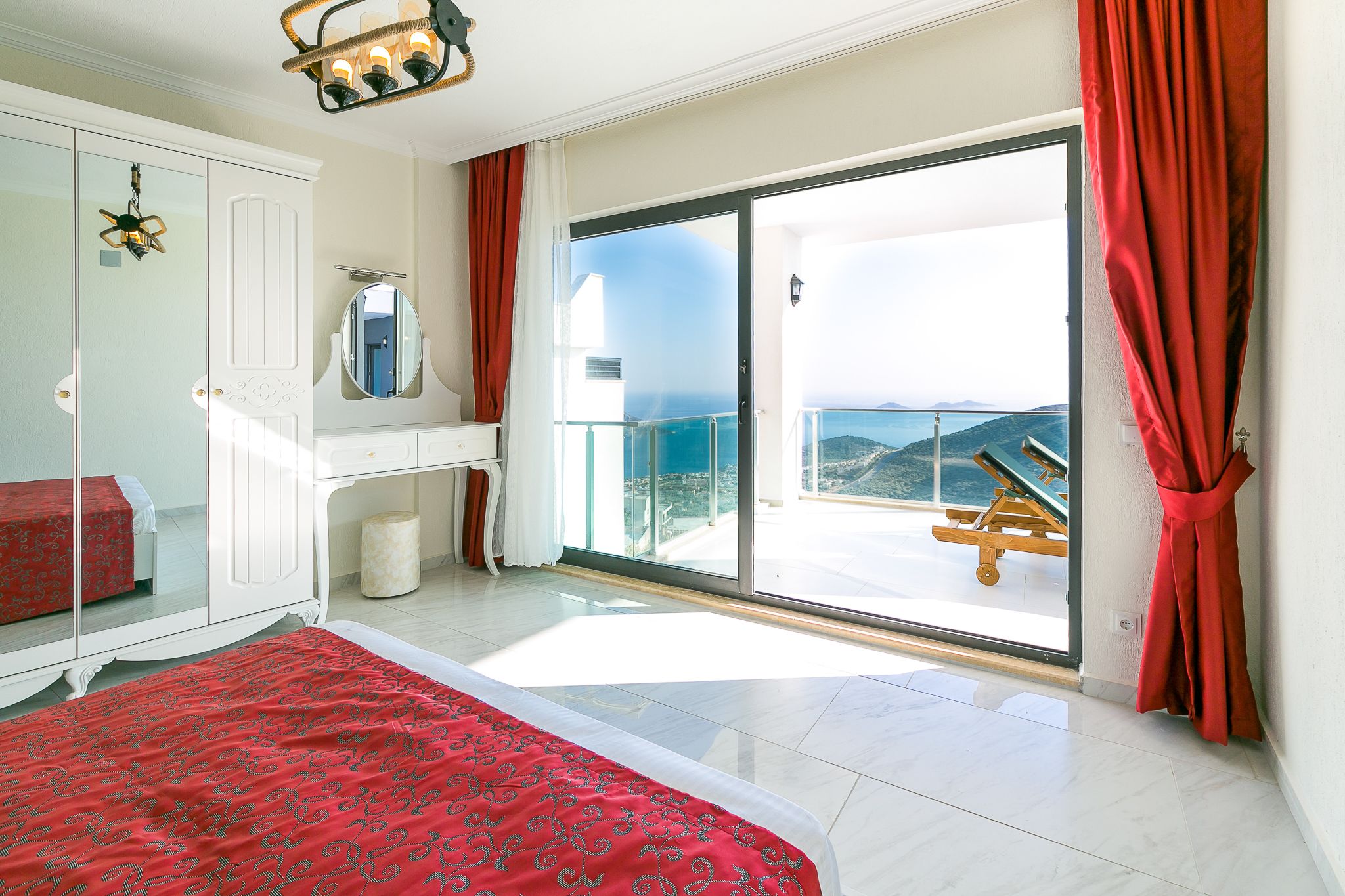 A bedroom with a large bed, a wardrobe, a mirror, and a sliding glass door leading to a balcony with a view of the sea.