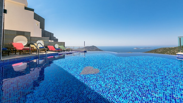 Une piscine à débordement aux carreaux de mosaïque bleus, des chaises longues rouges et une vue sur la mer et le paysage lointain.