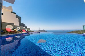 A infinity pool with blue mosaic tiles, red lounge chairs, and a view of the sea and distant land.