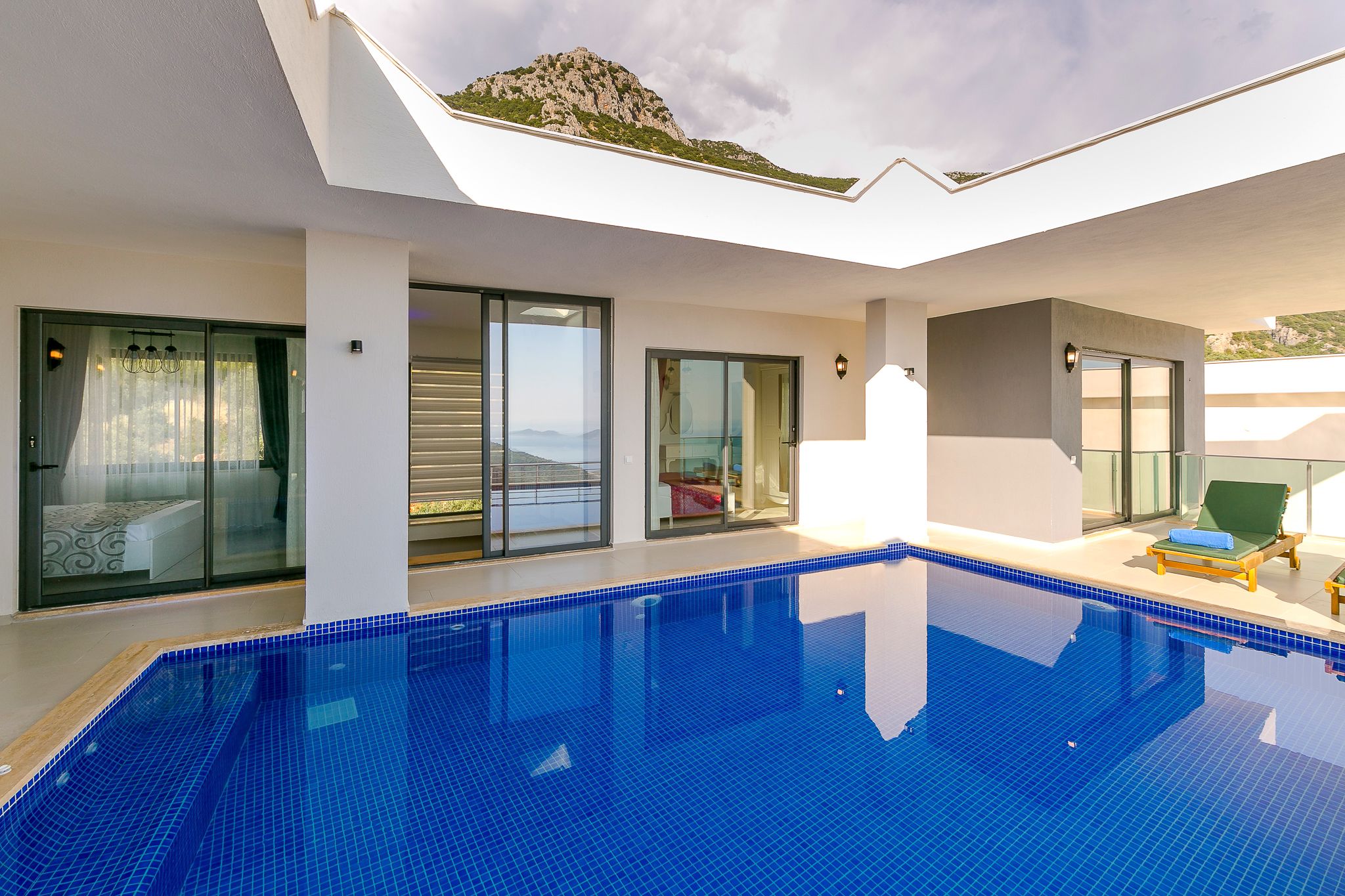 A modern indoor swimming pool with clear glass walls, a view of the mountains, and a sunlit lounge area.