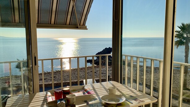 Een balkon met uitzicht op zee, een tafel met een bord, een kopje en een mok, en een raam met een strepenpatroon.