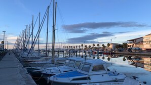 Een jachthaven met vele boten afgemeerd langs de waterkant, een heldere lucht en een gebouw met balkons op de achtergrond.