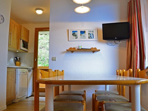 A compact kitchen with wooden cabinets, a microwave, and a refrigerator. There is a dining table with four chairs and a flat-screen TV mounted on the wall.