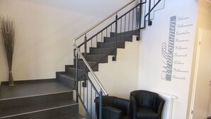 A staircase with black railings and dark tiled steps, leading to a landing area with two black chairs and a decorative wall with multilingual welcome signs. - Köhncke's Landgasthof und Hotel (Wiefelstede)