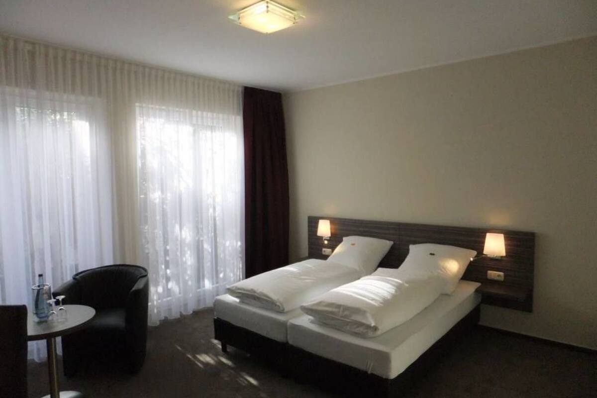A hotel room with a double bed, two bedside tables with lamps, a chair, and a small table with a bottle and glasses.