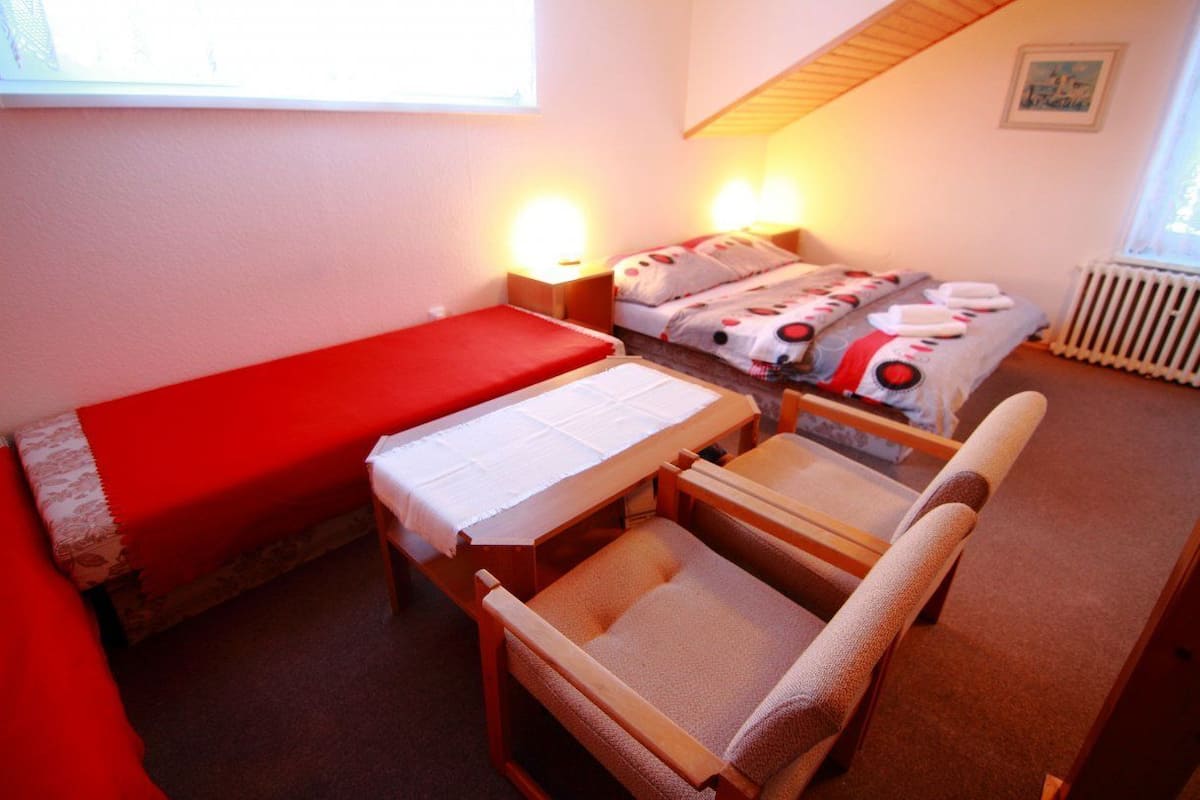 Una habitación pequeña y acogedora con un sofá rojo, una mesa de madera con mantel blanco y una cama con ropa de cama estampada. Hay dos lámparas de noche y un radiador en la pared.