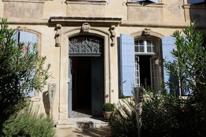 A building with a central entrance, flanked by two windows with blue shutters, and a small potted plant at the entrance. - Very comfortable independent studio of 30 m2, in a 19th century presbytery. (Tarascon)