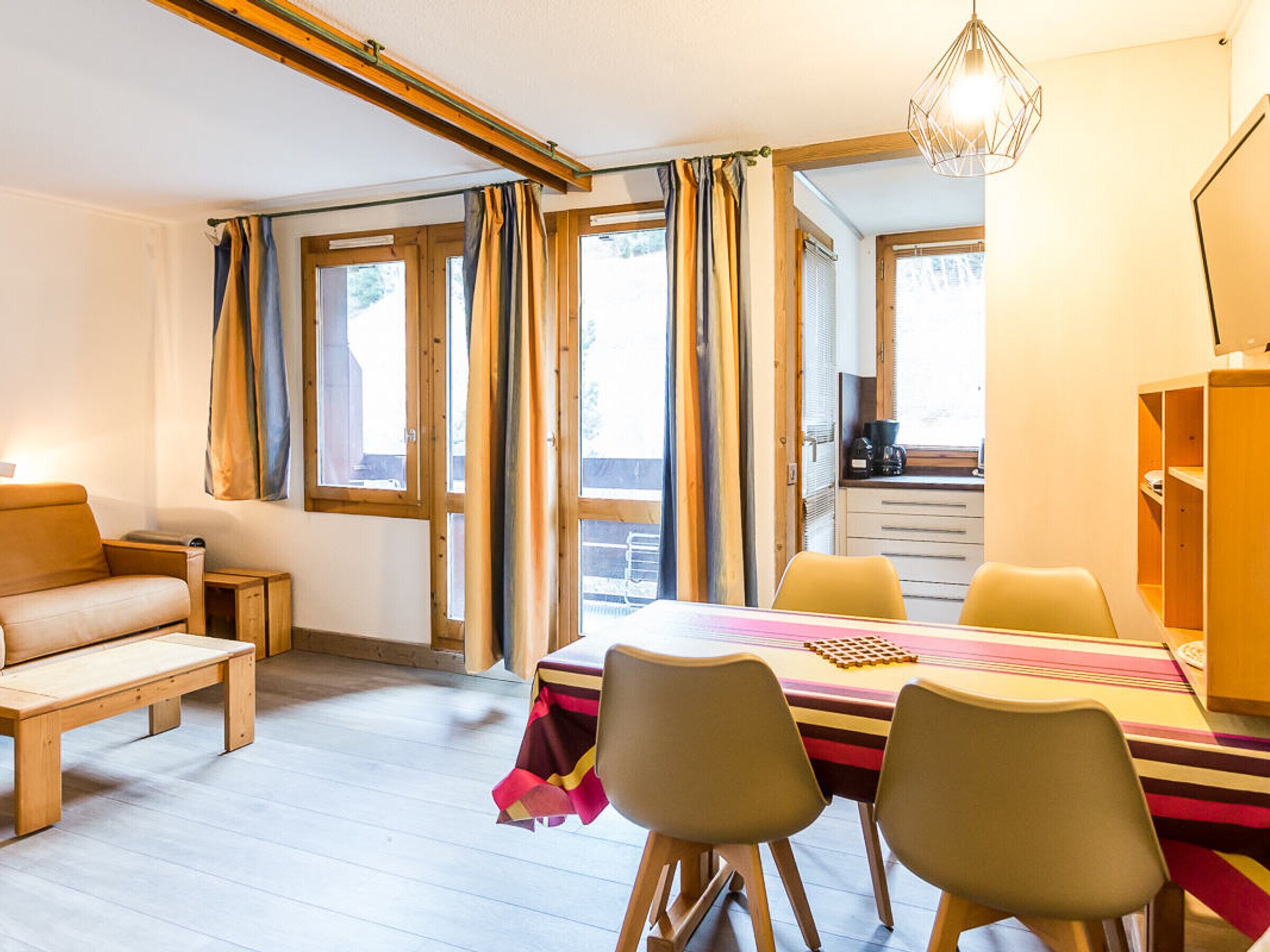 A cozy room with a wooden bench, a table with chairs, a desk, and a view through large windows.