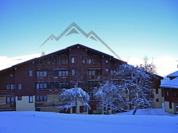 A multi-story building with a red facade and balconies, surrounded by snow-covered trees.