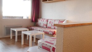 A cozy room with a sofa, two white tables, a window with curtains, and wooden shelves.